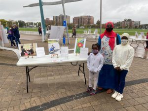 Alt= on the left of the image is a table with three photos sitting up on them. To the right of the table are three people standing and looking toward the camera. There is an adult and two children.