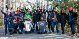 Group of people outdoors on a forest trail, dressed in winter clothing, some holding MSVU shirts, with trees and sunlight in the background.