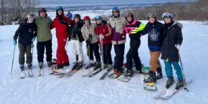 Group of people standing on a snowy slope wearing ski gear and helmets, each holding skis or snowboards, with a scenic winter landscape in the background.