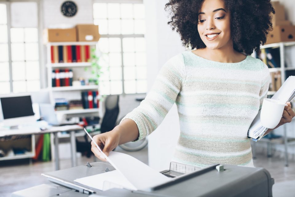 Smiling young woman at workplace. Holding folders with documents and cup, and using a copy machine. Tall windows with plants, filing cabinets with boxes and folders, computer and office seat as background.