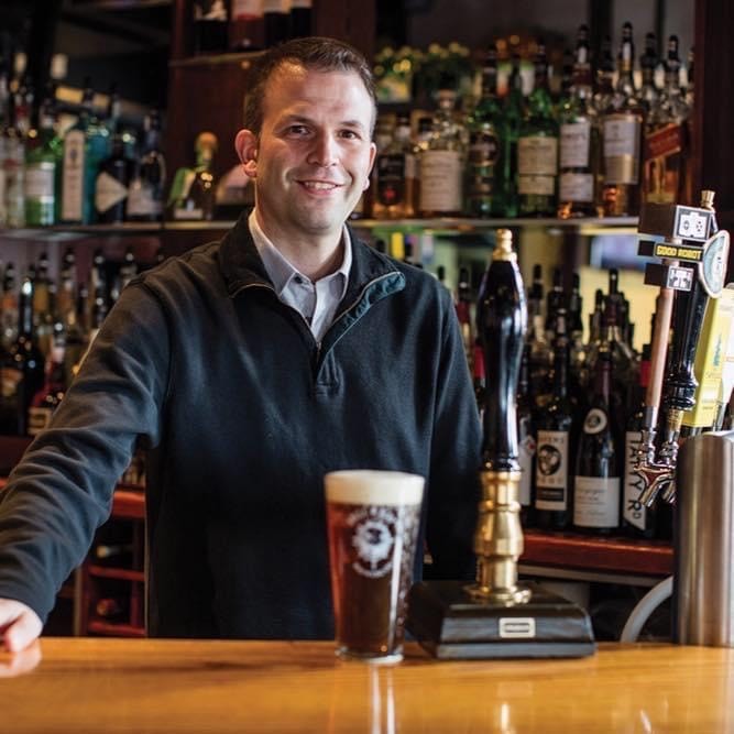 Adam Bower behind bar with glass of beer in front of him. Adam has short brown hair and is wearing a dark gray sweater with a pale gray button up shirt underneath. 
