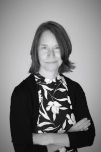 Photograph of Nancy Taber black and white. It shows a woman with dark chin-length hair with her arms crossed in front of her. She is smiling and looking directly at the camera.