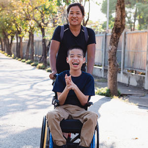 A caretaker with a child who is in a wheelchair, posing outside on a sidewalk