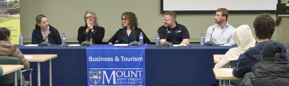 This photo shows five business and tourism career week panelists sitting behind a table with a blue table cloth on laughing during a panel session.
