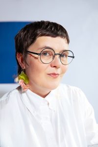 Blair Fornwald, wearing a white blouse and a green and peach coloured earring, along with black circular glasses