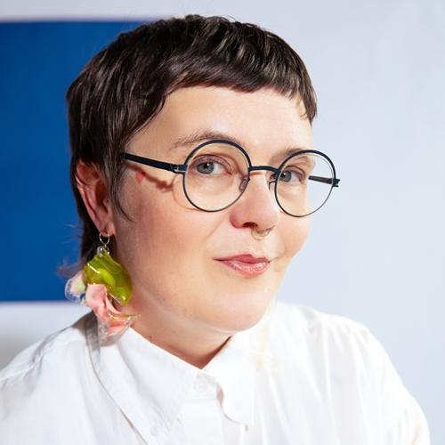 Blair Fornwald, wearing a white blouse and a green and peach coloured earring, along with black circular glasses