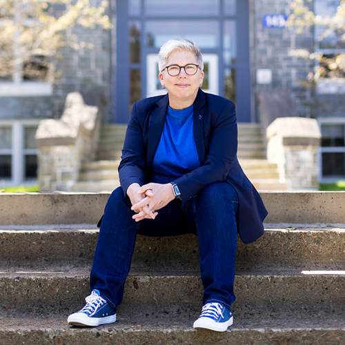 Dr. Joël Dickinson sitting on the steps in front of Evaristus. Wearing a navy blue blazer, blue sneakers, and a blue shirt.