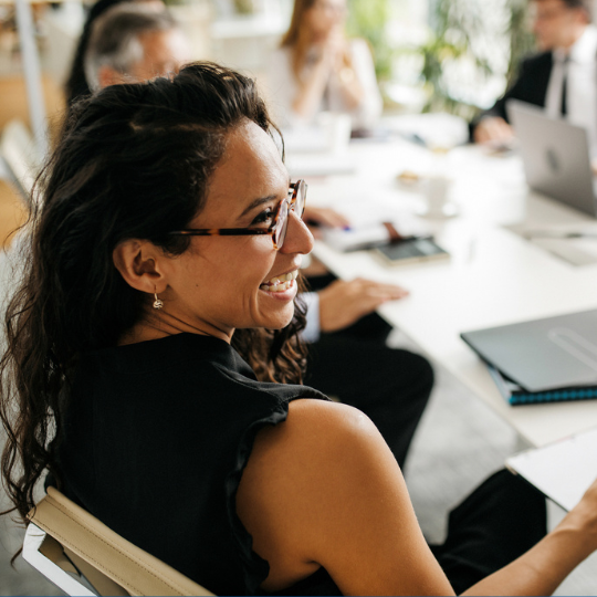 Smiling woman in glasses sits at a conference table holding a clipboard, engaged in a meeting with colleagues who are blurred in the background.