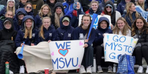 Several students watching a MSVU Mystics game outdoors, wearing MSVU Mystics clothing and holding signs that say, "We Love MSVU" and "Go Mystics Go!"