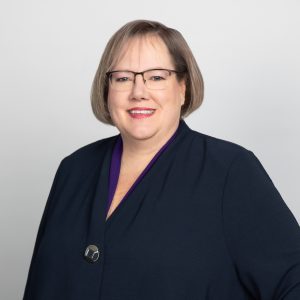 Darlene MacDonald MSVU Business and Tourism CEO panelist, 2025. Darlene is a white woman with short light brown hair and glasses. She is smiling and has a black suite jacket on. She is standing against a grey background.