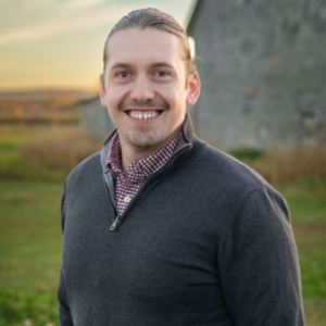 Joel Murphy, standing outdoors near a barn, wearing a grey sweater and smiling for the camera