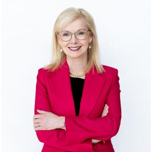 Joyce Carter MSVU Business and Tourism CEO panelist, 2025. Joyce is a white woman with blond hair. She is smiling and has a red suite with a black shirt. She is standing against a white background with her arms crossed in front of her