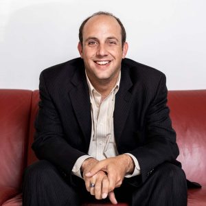 Mark Goldhar, MSVU Business and Tourism CEO panelist, 2025. Mark is a white man. He has a dark suit with a white buttoned shirt and is sitting on a red couch. Mark is smiling and is leaning forward with is elbows on his knees. His hands are clasped in front of him.