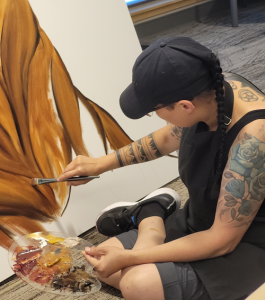 One of the artists of the Two-Eyed Seeing Mural, holding a paint brush and a palette, painting on a canvas