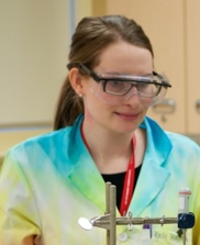 Dr. Kelly Resmer, wearing a rainbow tie-dyed lab coat with protective googles looking at lab equipment