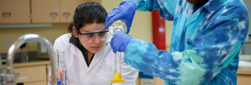 Two students, one wearing a white lab coat, the other wearing a blue lab coat, handling chemistry equipment in a lab
