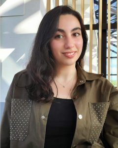 Hend Eldlk, wearing a brown jacket standing beside a window and smiling