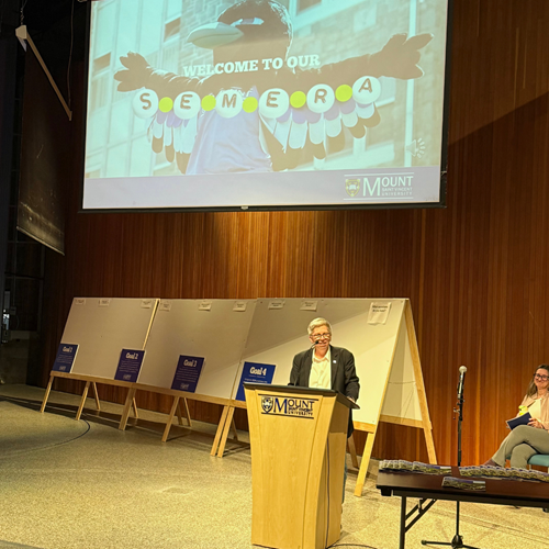 Dr Joël Dickinson speaking at the Strategic Enrolment Management (SEM) Launch, with the SEM Plan on a slideshow in the background