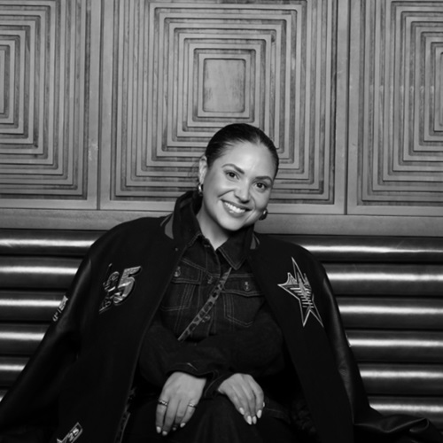 Bailey Williams, wearing a sports jacket, smiling at the camera. The photo has a greyscale filter applied to it.