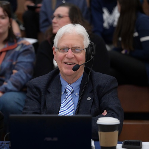 Dr. Robert Berard, wearing a suit and a headset, casting about the Basketball game