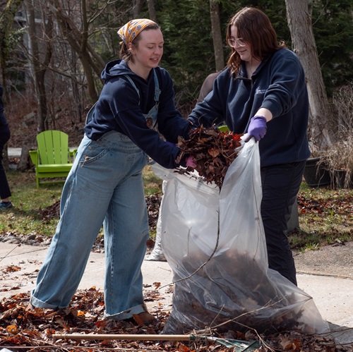 Two students putting leaves into a compostable trash bag outside of Assisi Hall