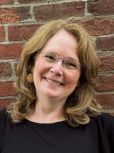Nathalie Blanchet, wearing a black sweater and glasses, smiling at the camera with a brick wall behind her.
