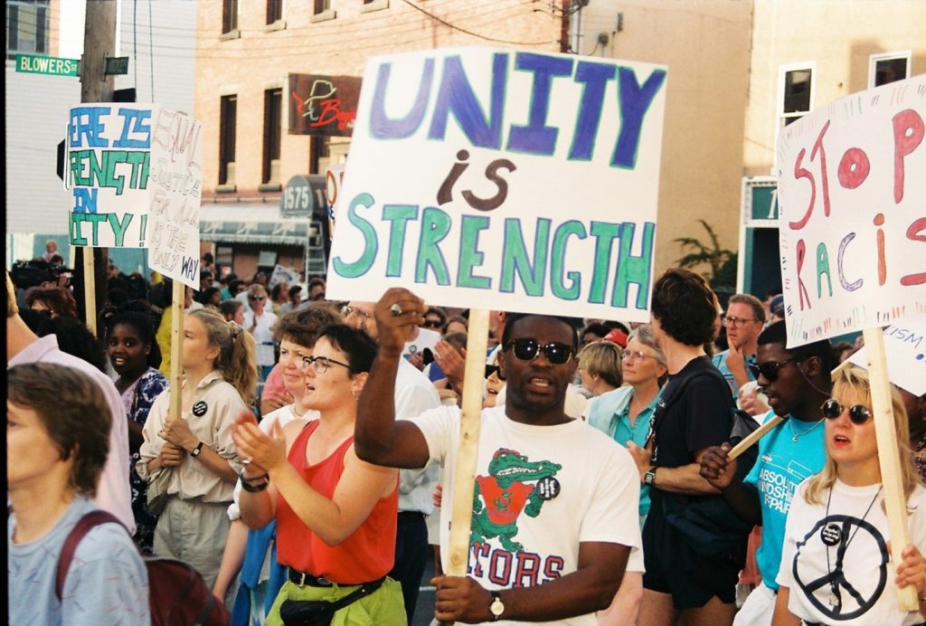 People protesting outside with a man wearing sunglasses and holding a sign that reads "Unity is strength" in the center of the photo