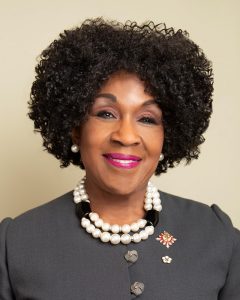 The Honourable Mayann Francis, wearing a grey blouse and a pearl necklace and earrings, smiling at the camera