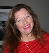 Catherine Martin, CM, wearing a red sweater in front of a painting, smiling at the camera with her head slightly tilted