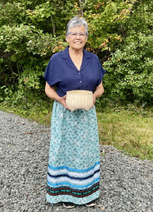 Della Maguire, holding one of the woven baskets. Della is outdoors and smiling at the camera.